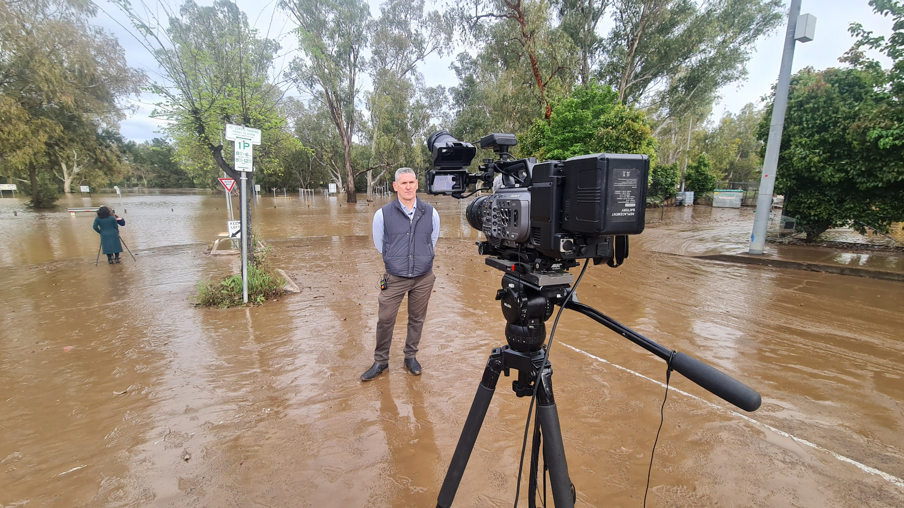 Evan Zell standing in floodwater beside a broadcast camera on a live cross