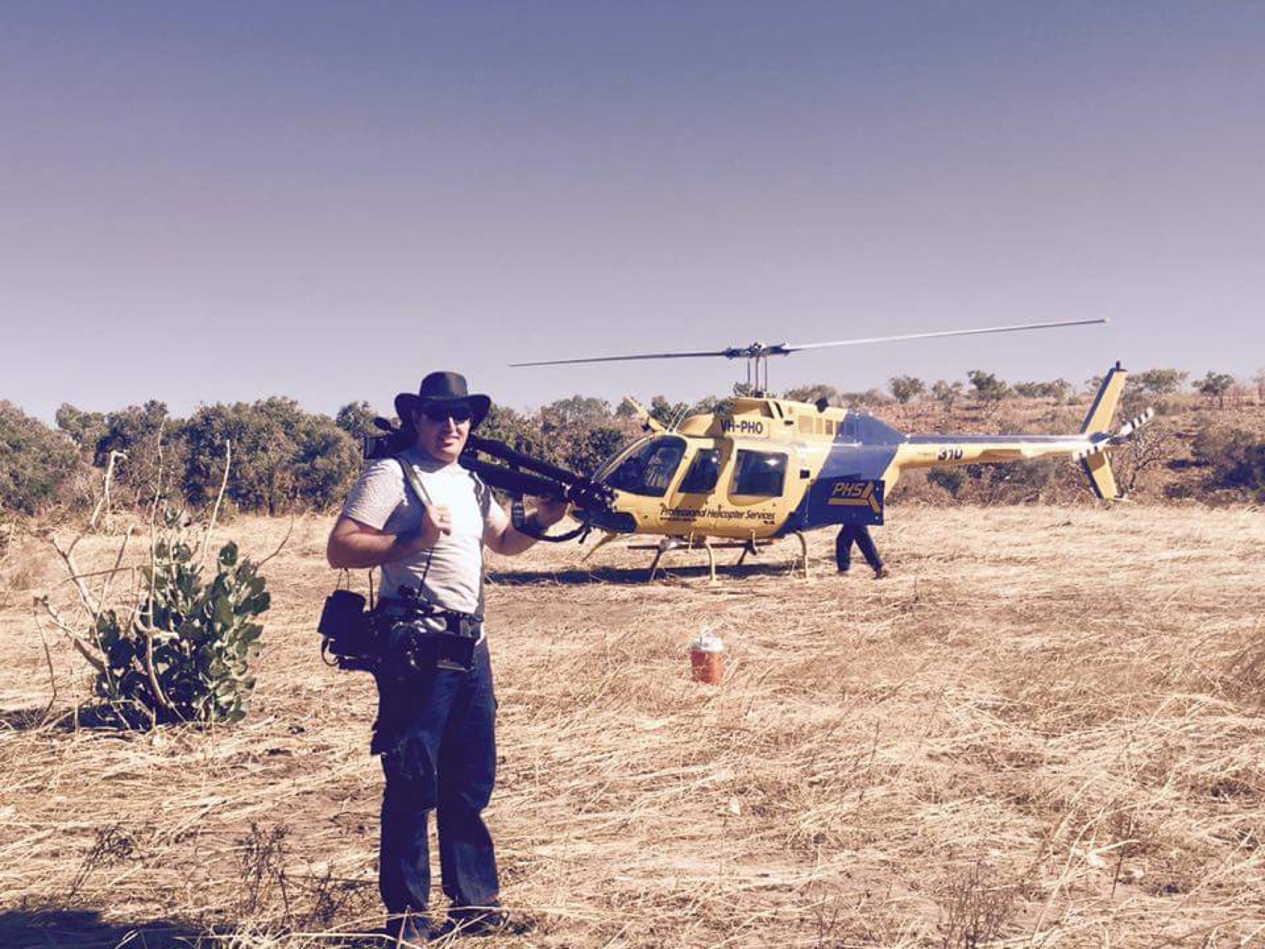 Evan with a camera in a field beside a helicopter