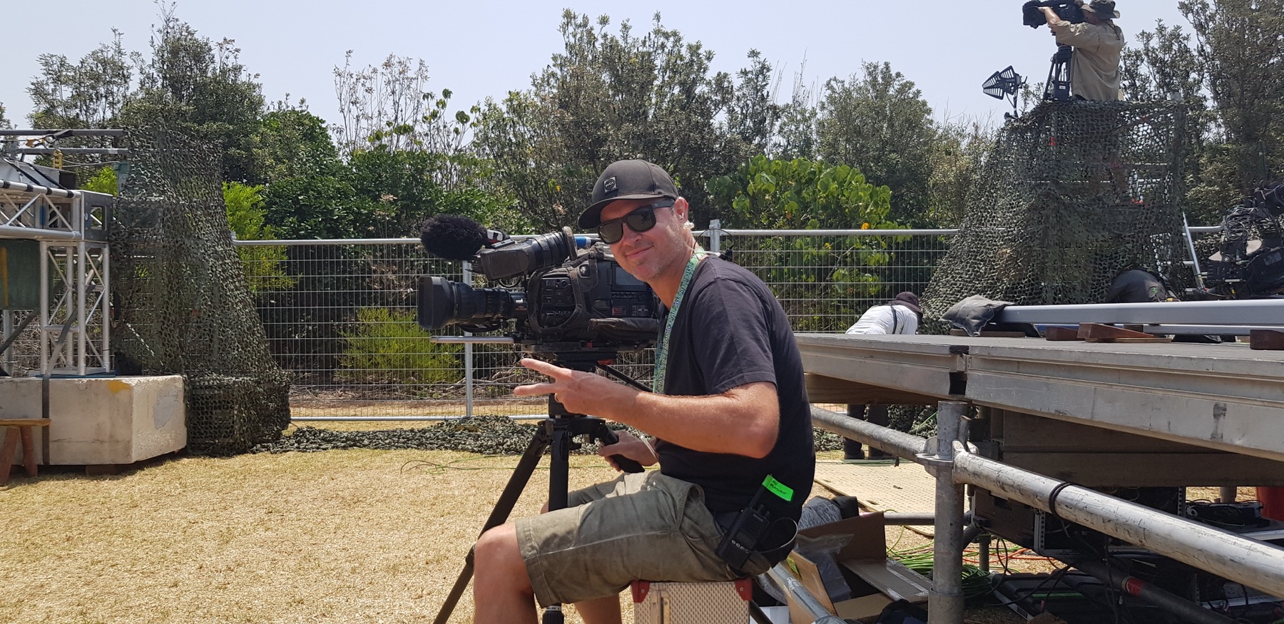 Evan sitting on a hillside with a camera, cap and sunglasses