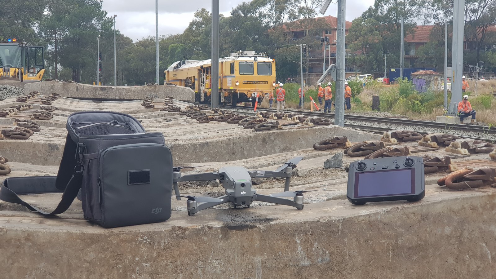 A drone on the ballast beside live rail track with a track machine and crew behind